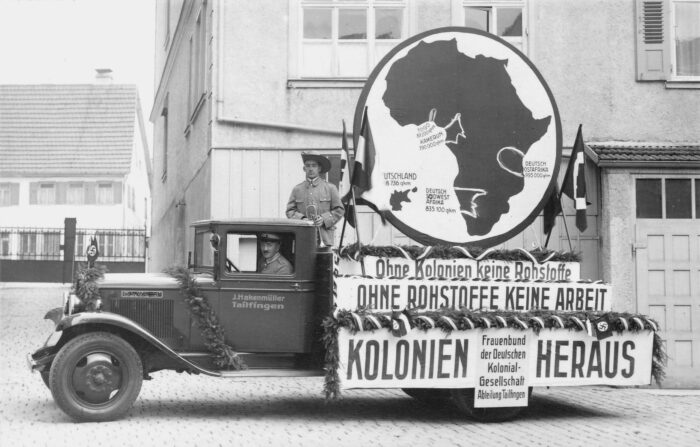 Historisches bild von einem Werbewagen mit, auf dem hinten eine große Afrika-Karte verzeichnet ist, mit den ehemaligen deutschen Kolonien. Darunter die Aufschrift "Ohne Kolonien keine Rohstoffe, ohne Rohstoffe keine Arbeit, Kolonien heraus"