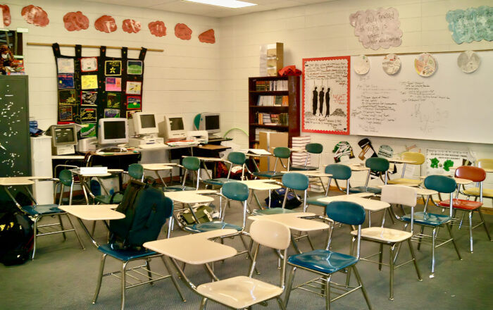 Ein altes Klassenzimmer mit leeren Stühlen und Tischen. Hinten im Bild sind alte Computer, an der Wand hängt Lernmaterial.