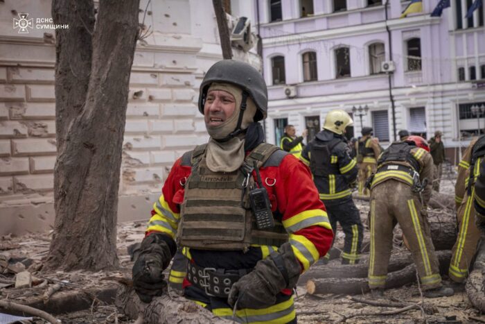 Ein Feuerwehrmann oder Katastrophenhelfer mit Helm und kugelsicherer Weste steht mit verzweifeltem Gesichtsausdruck auf einer von Schutt und Baumresten bedeckten Straße