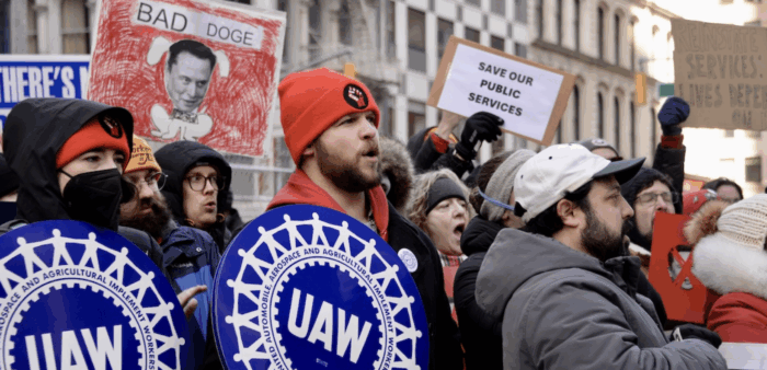 Zu sehen sind mehrere Leute, in dicken Jacken und mit Mützen, die Schilder halten, auf denen unter anderem UAW (der Name einer Gewerkschaft) steht. Auf einem Schild steht: Save our public Services.