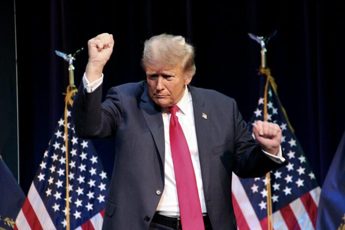 Das Bild zeigt US-Präsident Donald Trump. der auf einer Bühne zwischen zwei USA Flaggen steht und gerade zu tanzen scheint.
