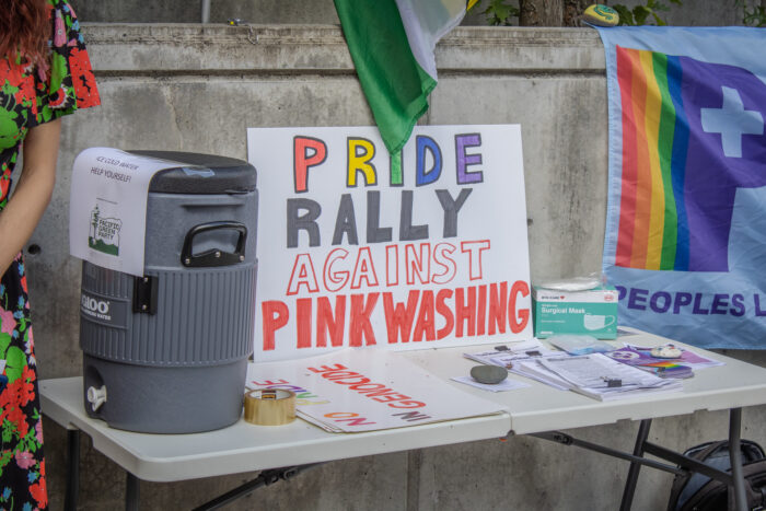 Ein Plakat mit der Aufschrift "Pride Rally against Pinkwashing" ist auf einem Tisch aufgestellt, daneben eine Packung Coronamasken, ein Wasserbehälter, Flyer und Pride-Sticker.