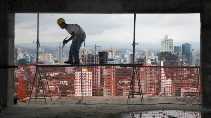 Ein Arbeiter balanciert in großer Höhe auf einem Holzgerüst, umrahmt von einer Öffnung für eine Fensterfront. Im Hintergrund die Skyline von Panama Stadt.
