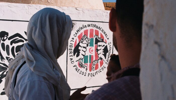drei Menschen unterhalten sich vor eine Wand mit Murals. Auf einem ist eine Hand zu sehen, die Ketten bricht, dahinter die Flagge von Westsahara. Drumherum die Beschreibung der internationalen Kampagne für Westsahara.