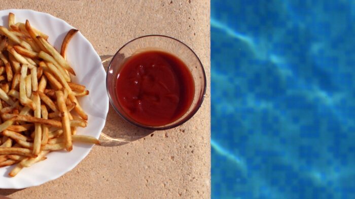 Eine Portion Pommes liegt auf einem weißen, am Rand gewellten Teller. Der Teller steht ganz nah am Beckenrand eines Freibades. Zwischen Wasser und Pommes befindet sich ein rundes kleines Glasschüsselchen mit einer Portion Tomatenketchup.