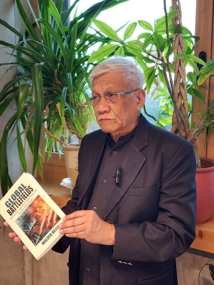 Walden Bello steht vor einer Fensterbank auf der einige Zimmerpflanzen stehen. Er trägt ein schwarzes Hemd, einen schwarzen Anzug und eine randlose Brille. Seine Haare sind grau. Er schaut ernst. In der Hand hält er sein Buch Global Battlefields.