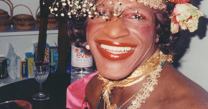 Nahaufnahme von Marsha P. Johnson. Sie lacht in die Kamera. Sie trägt roten, glänzenden Lippenstift und Rouge, sowie Lidschatten in Rosetönen. In den glänzenden lockigen Haaren hat sie Blumen gesteckt. Etwas Schleierkraut ragt über die Augenpartie, fast wie ein echter Schleier. Um den Hals trägt sie mehrere goldene Bänder, die an Schleifenband von Geschenken erinnern. Sie trägt eine pinke Satinbluse mit Carmenausschnitt und Perlenohrringe. Im Hintergrund ist ein Regal zu sehen, darauf ein halbgefülltes kristall Stilglas und eine Evian-Wasserflasche.