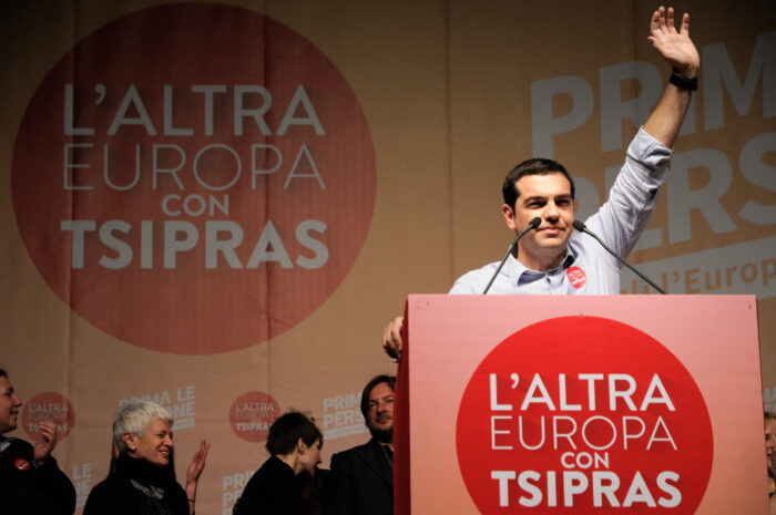 Man sieht Alexis Tsipras winkend an einem Rednerpult stehen.