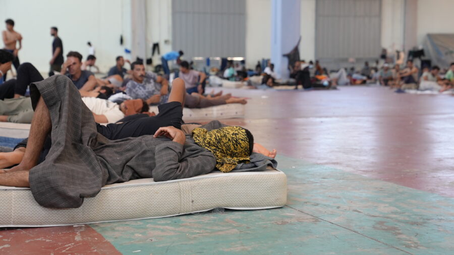 In einer großen Halle liegen und sitze viele Männer aus einigen Matratzen oder auf dem Boden. Sie verbergen ihre Gesichter mit den Händen oder unter Tüchern.