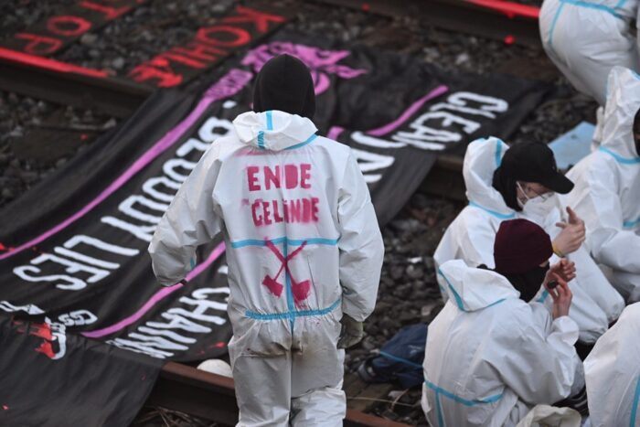 Mittig eine Person in weißem Overall mit dem Rücken zur Kamera. Auf dem Overall die Inschrift "Ende Gelände", vor ihr Bahnschienen mit Transparent, rechts weitere Personen in Overalls auf den Schienen hockend.