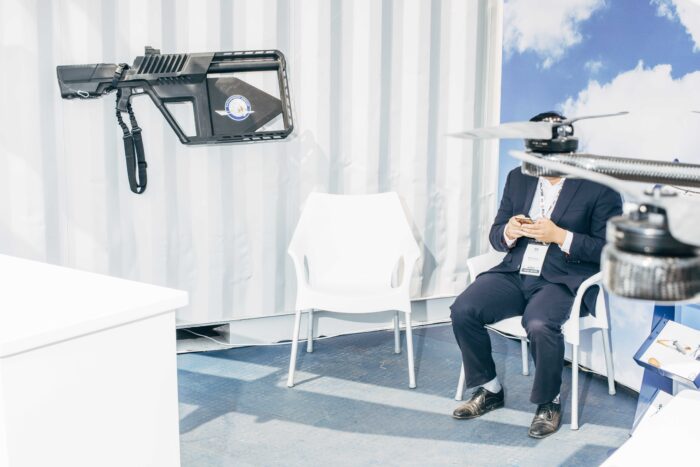 Ein Mann im Anzug sitzt auf einem Messestand und tippt in sein Telefon, das Gesicht ist verdeckt von einem technischen Gerät, an der Wand hängt eine Schusswaffe