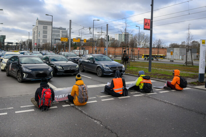 Fünf Aktivist*innen der Letzten Generation sitzen mit Warnwesten in einer Reihe auf der Straße, vor ihnen Autos.