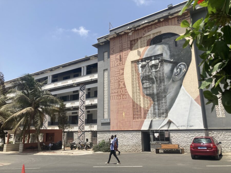 Bild eines Murals an der Wand der Universität. Darauf zu sehen ist ein Porträt von Cheikh Anta Diop, mit Brille und Schnautzer, der in die Ferne schaut. Um seinen Kopf ist eine Art Heiligenschein.