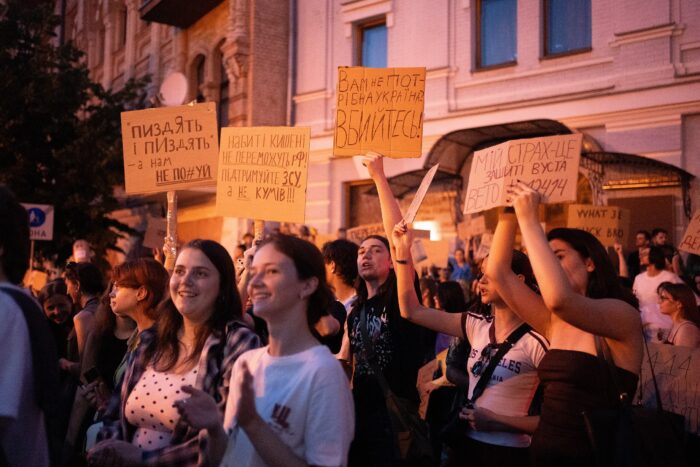 Junge Demonstrant*innen mit Pappschildern, auf denen ukrainische Protestsprüche stehen, im Abendlicht