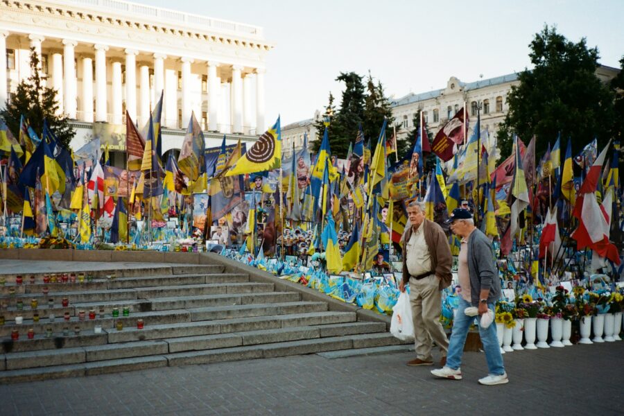 Zwei alte Männer gehen an einem Platz mit vielen blau-gelben Fahnen und Wimpeln und Fotos von Soldaten vorbei, auf der linken Bildseite ist eine Treppe zu sehen, auf der Kerzen stehen