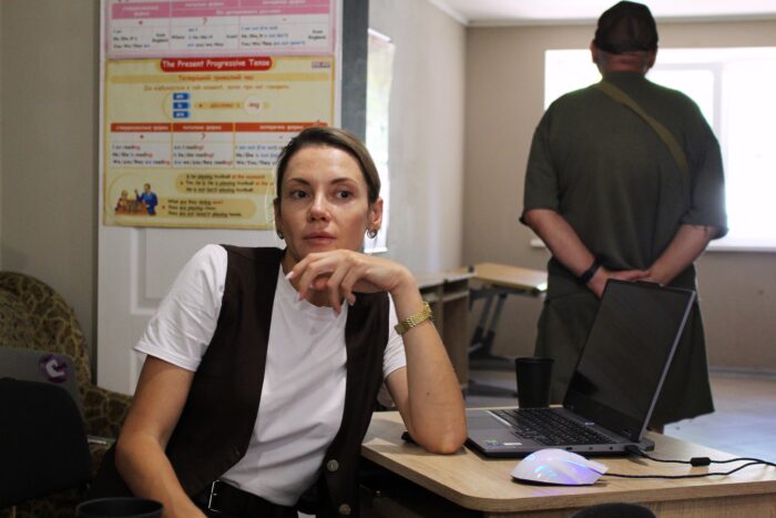 Eine Frau sitzt an einem Tisch mit Computer, hinter ihr steht ein Mann, der dem Betrachter den rücken zuwendet und mit verschränkten Armen in die Ferne schaut.