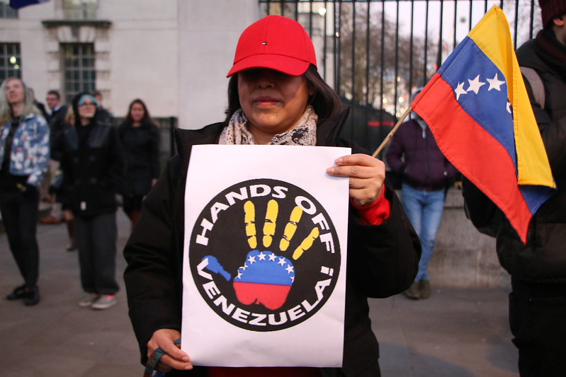Auf dem Bild ist eine Person mit langen dunklen Haaren und einer roten Cap zu sehen, die ein Plakat hält: Hands off Venezuela steht da drauf. Außerdem schwenkt die Person eine kleine Venezuela-Flagge