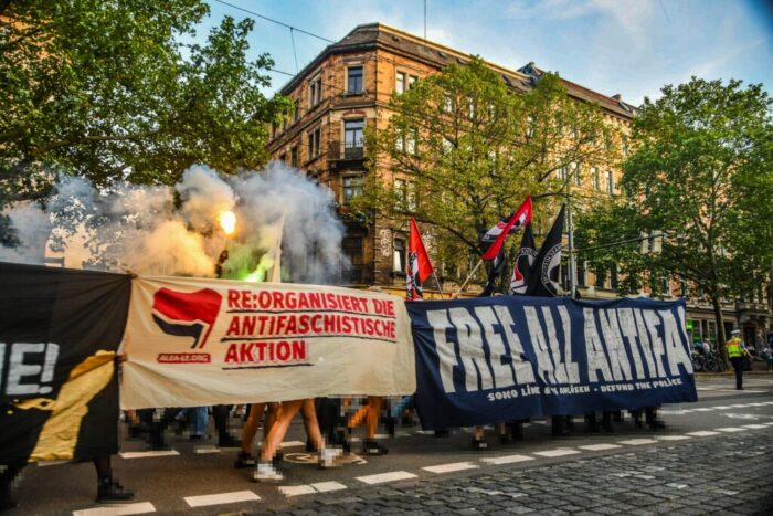 Zu sehen sind drei Demobanner mit verschiedenen Slogans, beispielsweise „Free all Antifas”, sowie Fahnen der Antifa. Unten sind die Füße und Beine der Demo-Teilnehmer*innen zu erkennen.