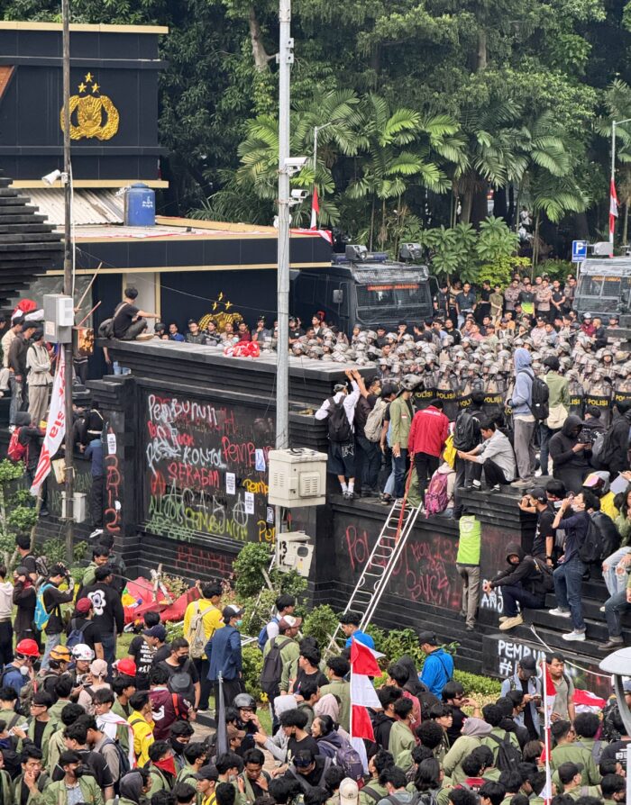 Eine große Gruppe junger Menschen steht vor einer besprühten Mauer, einige sind hinaufgeklettert, dahinter steht eine sehr große Gruppe Polizisten mit Helmen und ein Wasserwerfer