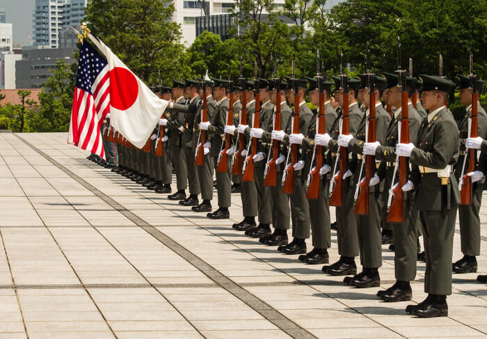 Zu sehen ist eine Militärparade, anlässlich des Besuchs US-amerikanischer Befehlshaber in Japan. Die uniformierten Soldaten stehen in einer geraden Reihe und halten eine japanische und eine US-amerikanische Flagge in der Hand