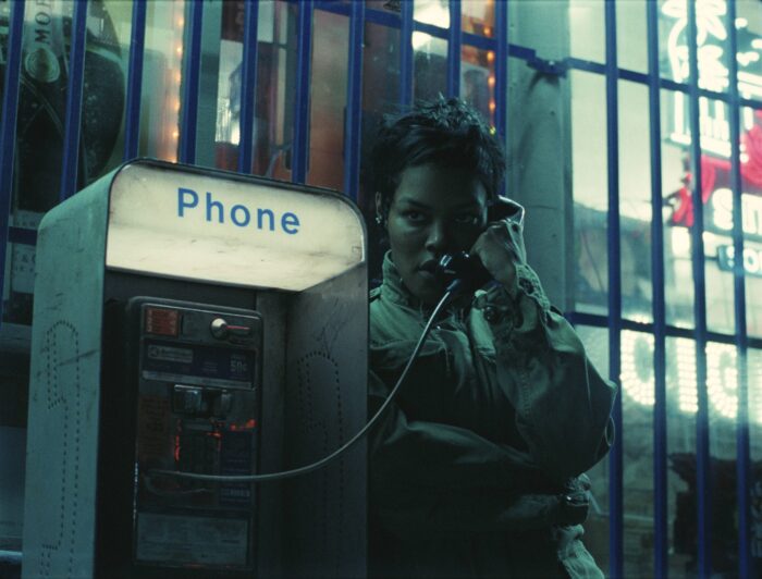 Teyana Taylor, mit Army Jacke, lehnt nachts an einer altmodischen Telefonzelle und telefoniert.