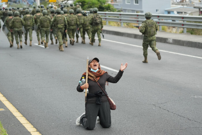 Bild einer Frau, die weinend auf einer Straße kniet und klagt, eine Hand nach oben streckend. Hinter ihr läuft eine Gruppe von Soldaten von ihr weg.