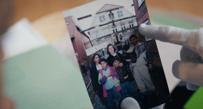 Ein Finger in Handschuhen zeigt auf eine Fotografie, darauf eine Familie mit Kindern, im Hintergrund ein Haus