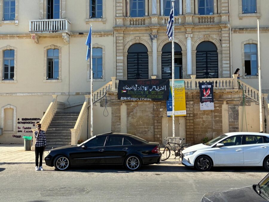 Das Bild zeigt ein Gerichtsgebäude auf Kreta. Davor packen zwei Autos. Eine Person steht vor der Treppe, die ins Gebäude führt, und schaut richtung Kamera