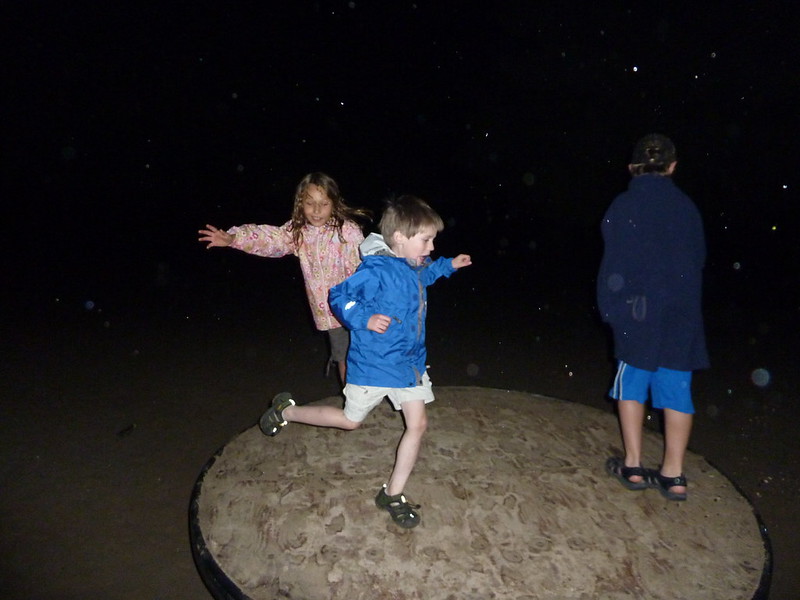 Drei Kinder spielen rennen auf einer Drehscheibe. Das Bild ist im Dunkeln aufgenommen, die Kinder sind vom Blitz erleuchtet und haben offensichtlich Spaß