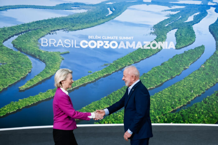 Im Hintergrund ein großes Bild vom Regenwald, davor Ursula von der Leyen und Lula, die sich die Hände reichen.