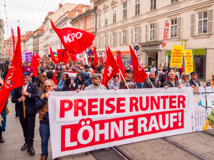 Die Spitze eines Demozuges mit einem Banner, auf dem steht "Preise runter, Löhne Rauf"; mehrere Rote Fahnen mit dem Kürzel "KPÖ".