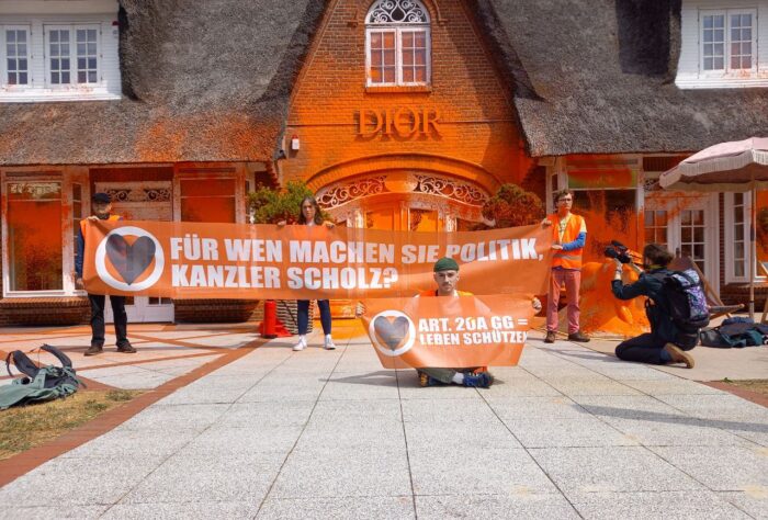 Aktivist*innen halten ein Banner mit der Aufschrift "Für wen machen Sie Politik, Kanzler Scholz?", im Hintergrund eine gediegene Villa mit Reetdach, das mit orangener Farbe besprüht ist.