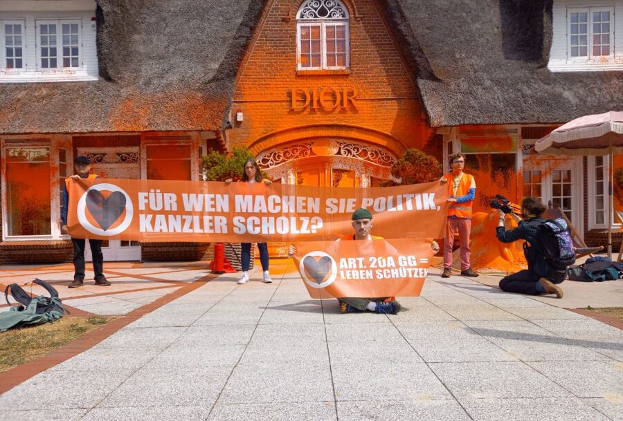 Aktivist*innen halten ein Banner mit der Aufschrift "Für wen machen Sie Politik, Kanzler Scholz?", im Hintergrund eine gediegene Villa mit Reetdach, das mit orangener Farbe besprüht ist.