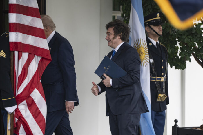Milei läuft hinter Trump her in den Eingang in eine Gebäude und zeigt zwei Daumen hoch. Zwei Soldaten halten jeweils die Fahnen von Argentinien und den USA.
