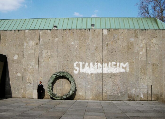 Kunstwerk "Stammheim" von Olaf Metzel: Ein übergroßer Trauerkranz aus grünem Stahl lehnt an einer Betonwand, auf die das Wirt "Stammheim" gesprüht ist. Neben dem Kranz steht ein Mensch.