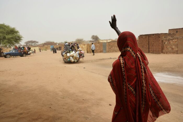 Eine Frau in einen roten Umhang steht auf einer Sandstraße und winkt einem abfahrenden Auto zu.