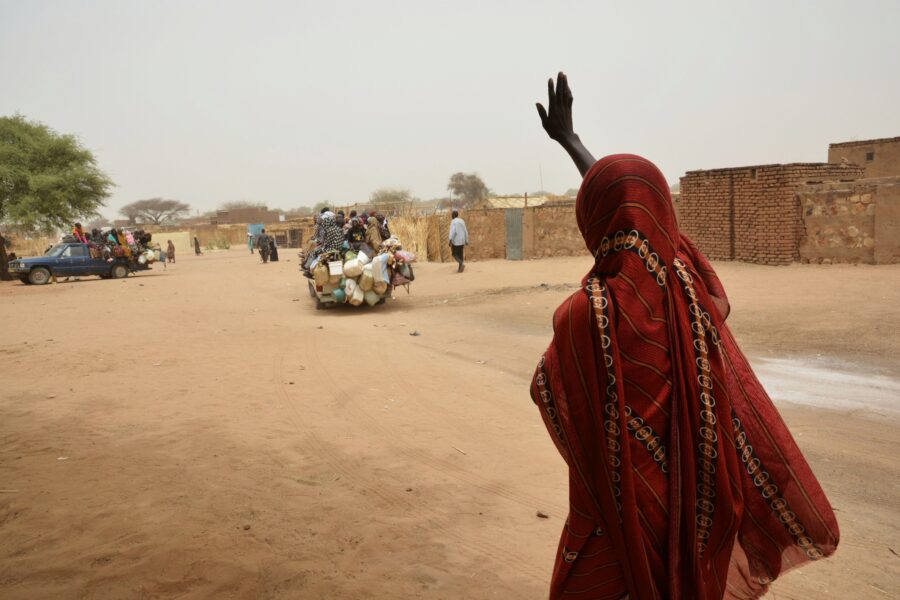Eine Frau in einen roten Umhang steht auf einer Sandstraße und winkt einem abfahrenden Auto zu.