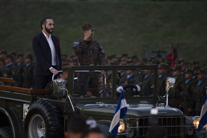 Foto einer Militärparade: Der Präsident steht auf der Ladefläche eines Militär-Geländewagens, neben ihm salutiert ein Offizier den Am Rand der Straße aufgestellten Soldaten.