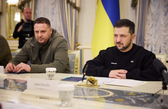 Andriy Yermak (links) und Volodymyr Selenskyj (rechts) sitzen an einem gold dekorierten Tisch, im Hintergrund ist die ukrainische Fahne zu sehen
