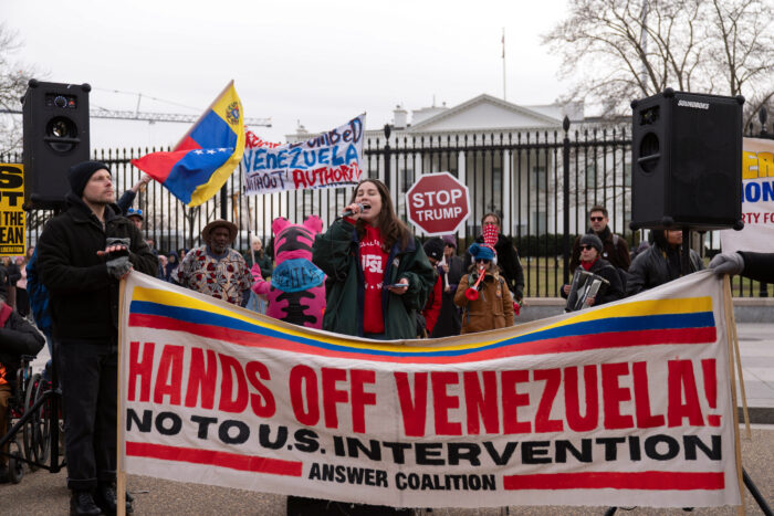 Protestierende nach dem US-Angriff auf Venezuela vor dem Weißen Haus. Eine Frau spricht ins Mikrofon, vor ihr ein Banner, auf dem Steht Hands off Venezuela (Hände weg von Venezuela).