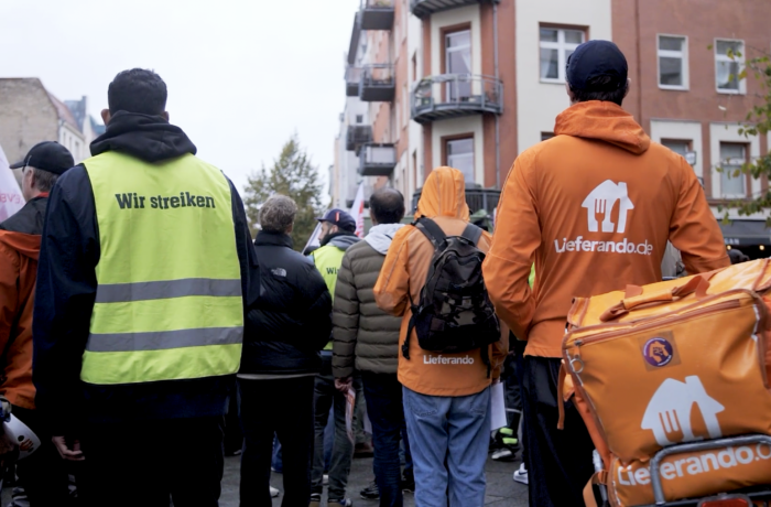 Mehrere Personen von hinten, die Person links im Bild trägt eine Weste mit der Aufschrift "Wir streiken", die Person rechts eine Lieferando-Jacke und lehnt an iner Lieferando Box auf einem Fahrrad