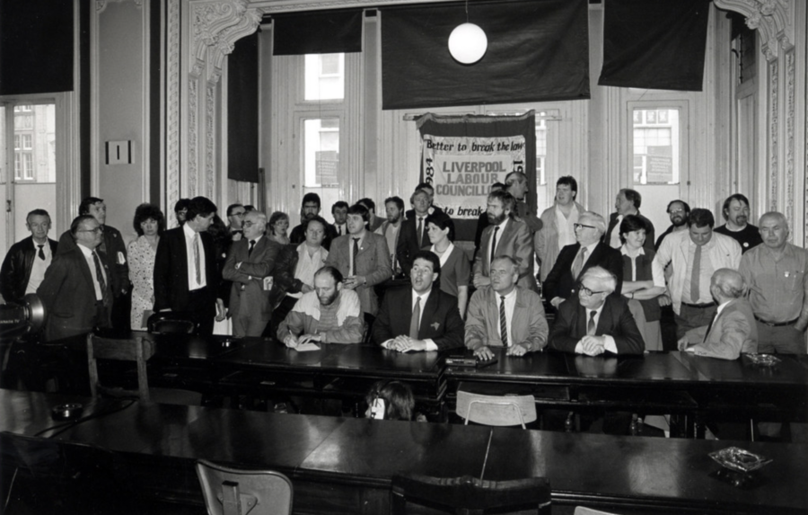 Man sieht auf einem Schwarzweißbild eine größere Menschengruppe in einem Saal, hinter ihnen hängt ein Transparent, auf dem steht Liverpool Labour Council.