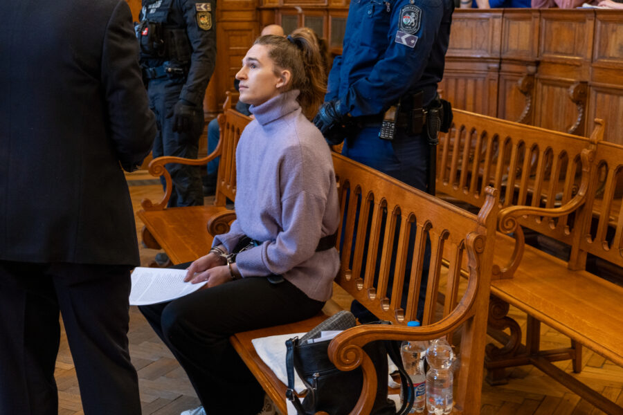 Man sieht eine Person in einem lila Pullover, die mit gefesselten Händen auf einer Gerichtsbank sitzt.