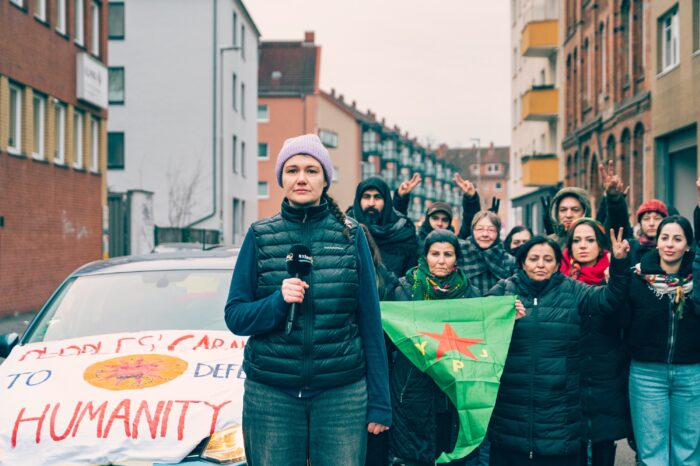 eine person mit Mikrofon steht vor einer Gruppe Frauen mit YPJ fahne und einem Auto, auf dessen Motorhaube ein Transpareent befestigt ist