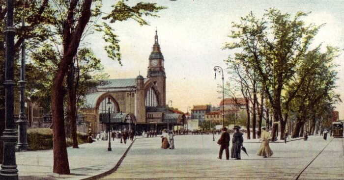 Hinten ein Gebäude mit Turm, davor kleine Personengruppen in der Mode zu Beginn des 20. Jahrhunderts daneben Bäume, Straßen und Gehwege.