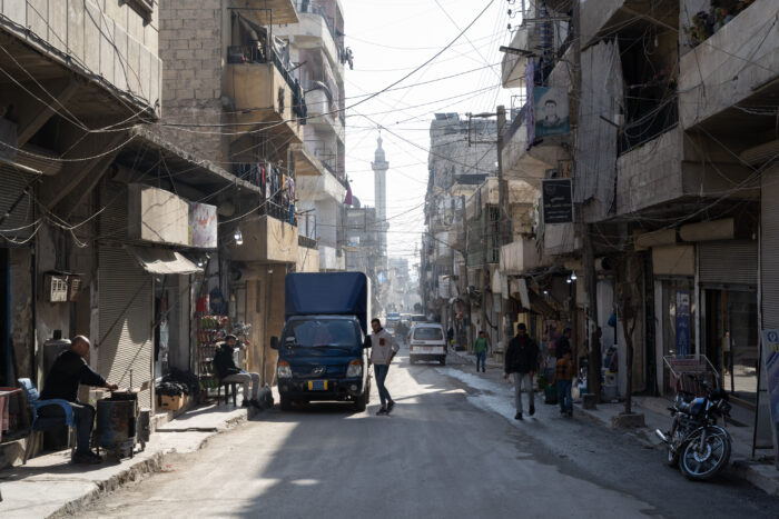Eine Straße im Stadtteil Sheikh Maqsoud im nordsyrischen Aleppo, in der Autos und einige Menschen stehen.