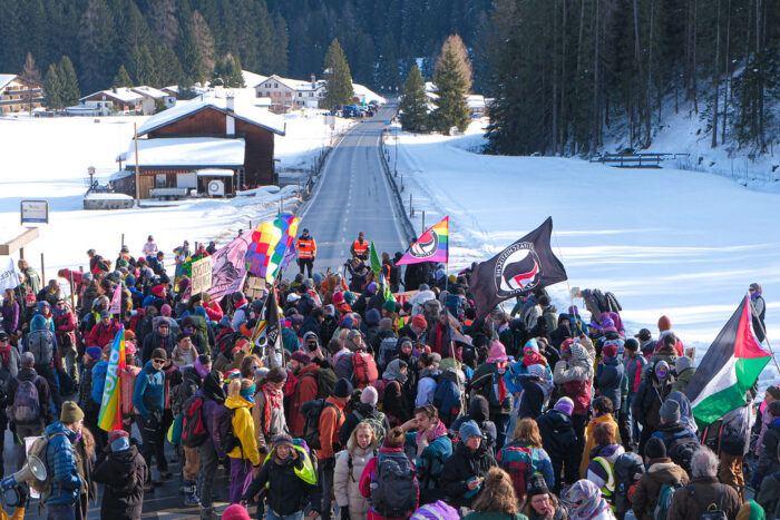 Demoaufnahme auf einer verschneiten Landschaft umgeben von Bergen treffen sich Demonstrierende. Die Polizei sperrt ihr den Weg.
