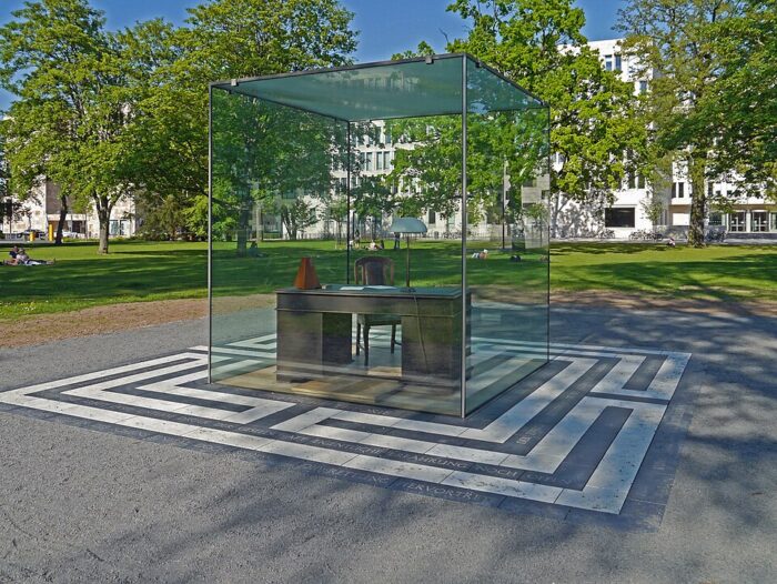 In einem Glaskasten steht ein Schreibtisch, dahinter Bäume und eine Rasenfläche.