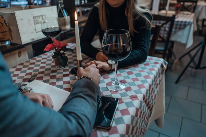 Das Bild zeigt offenbar eine Frau und einen Mann, die an einem Tisch in einem Restaurant sitzen und Händchen halten. Die Köpfe der beiden Personen sind auf dem Foto abgeschnitten. Neben ihnen steht Rotwein.