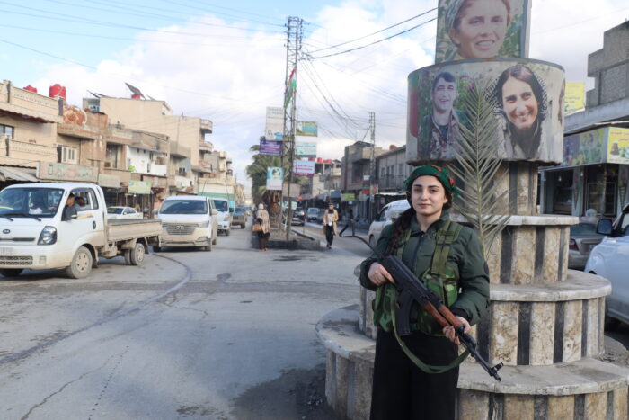 Das Bild zeigt eine junge Frau in der Uniform der kurdischen Frauenverteidigungseinheit YPJ. Sie trägt eine auf den Boden gerichtete Waffe in der Hand und lächelt in die Kamera